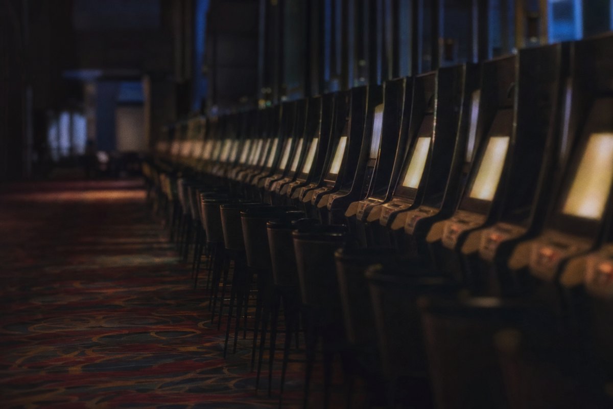 Lange Reihe von Spielautomaten in einem abgedunkelten Gang, die Bildschirme nur als helle Flächen erkennbar, davor leere Barhocker auf gemustertem Teppich