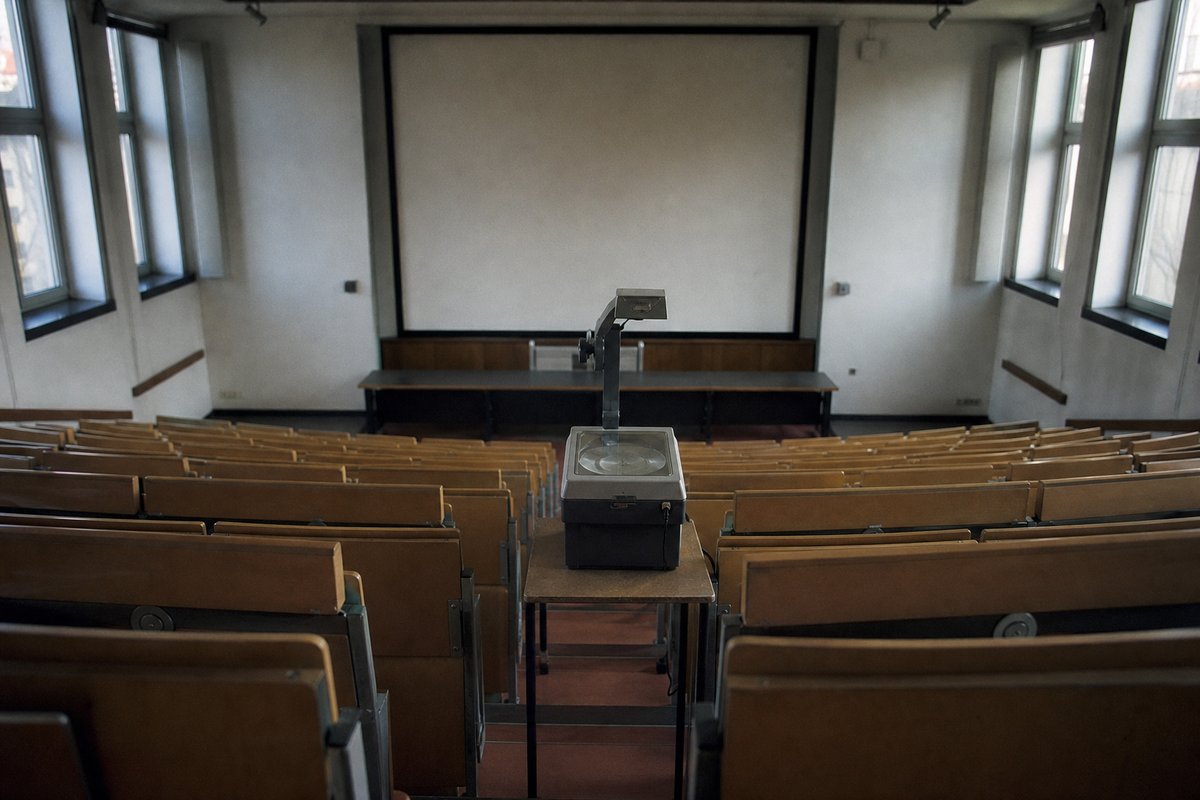 Blick von hinten in einen Hörsaal mit leeren Klappsitzen, Projektionsfläche und einem Overheadprojektor im Gang